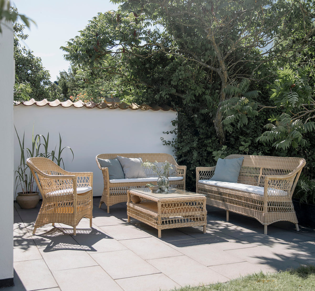 Dette er et livsstilsbillede af Charlot 2-personers sofa i natural ArtFibre Aluminium, på en kystterrasse med sivtag, havudsigt og stribede puder.