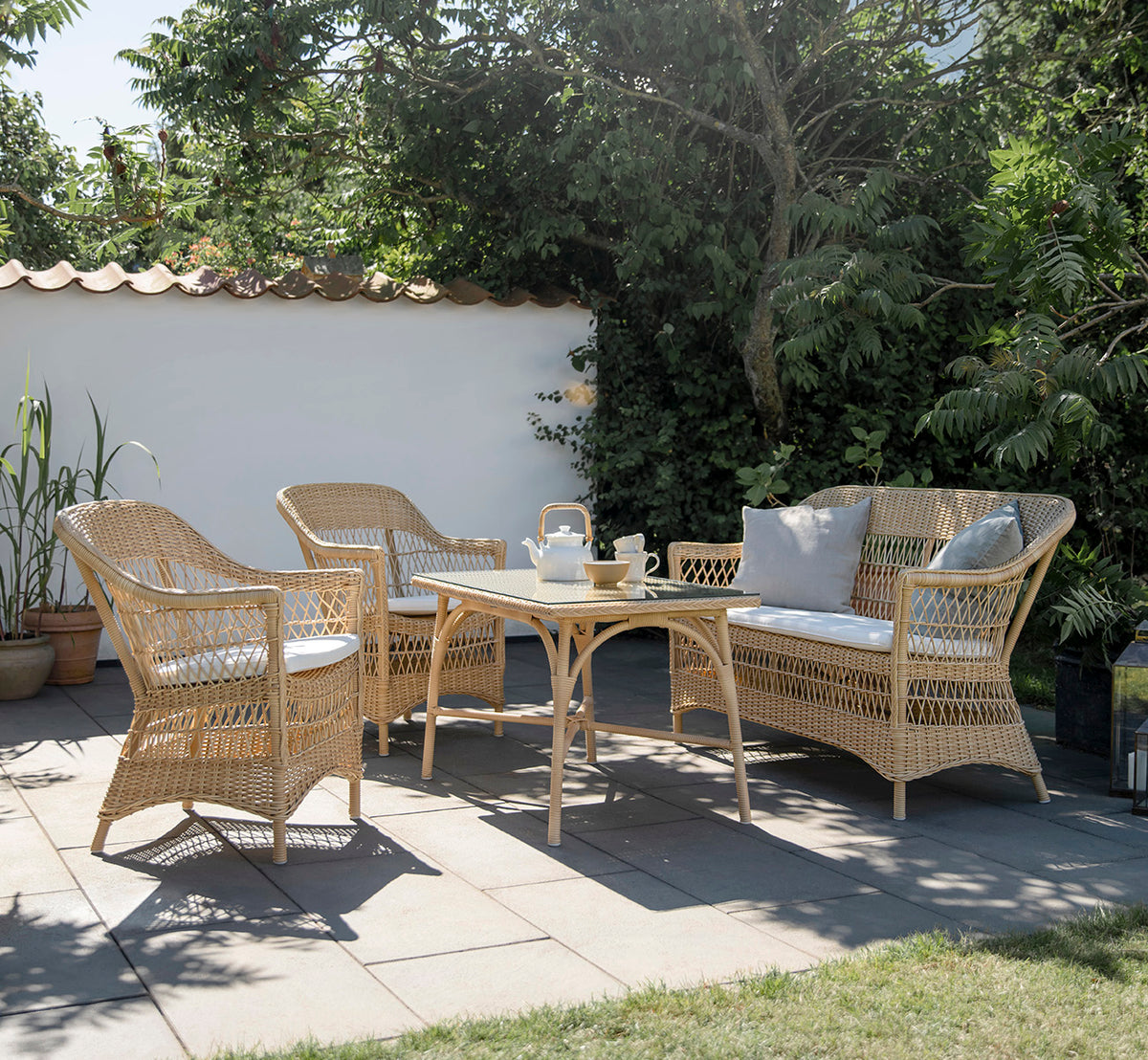 Dette er et livsstilsbillede af Charlot 2-personers sofa i natural ArtFibre Aluminium, på en kystterrasse med sivtag, havudsigt og stribede puder.
