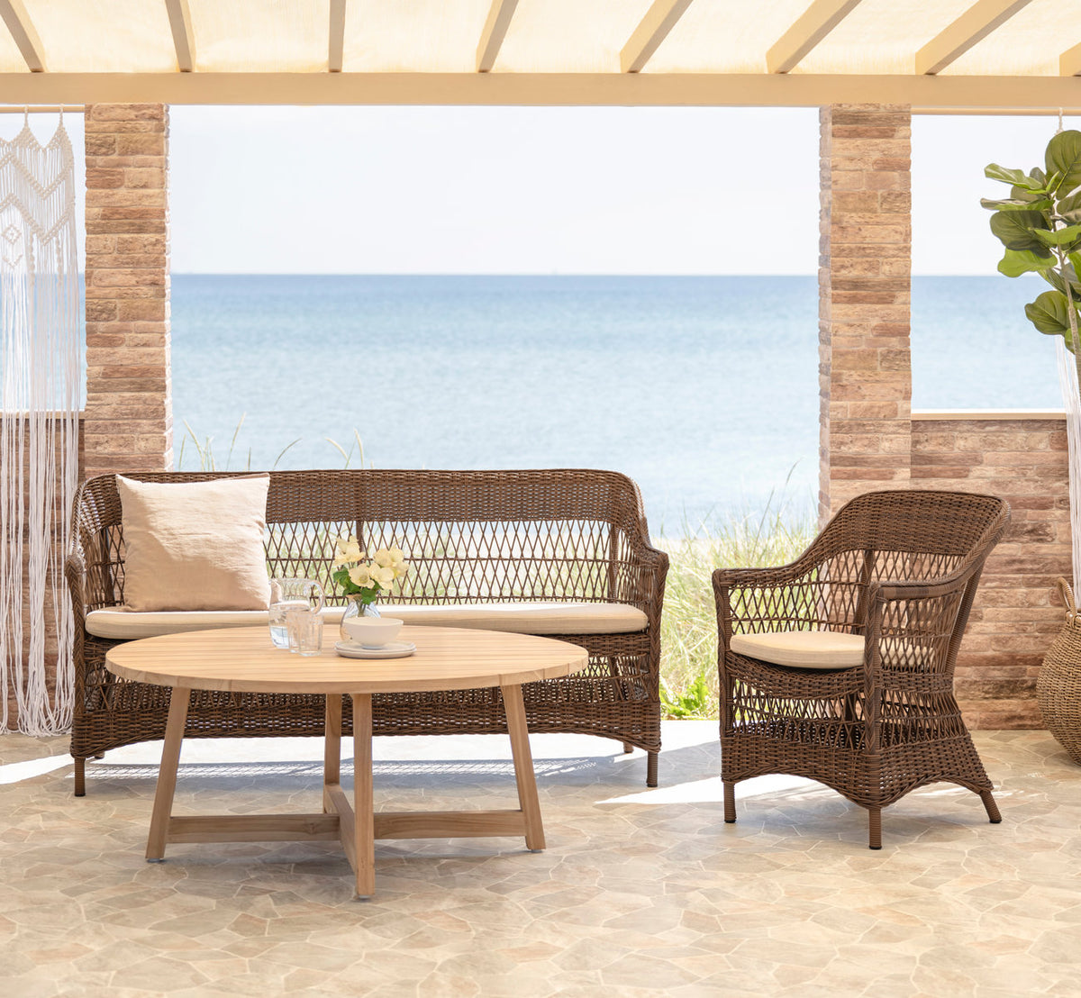 Dette er et livsstilsbillede af Charlot 3-personers udendørs sofa i brown artfibre aluminum. Med teak sofabord på en terrasse ved havet.
