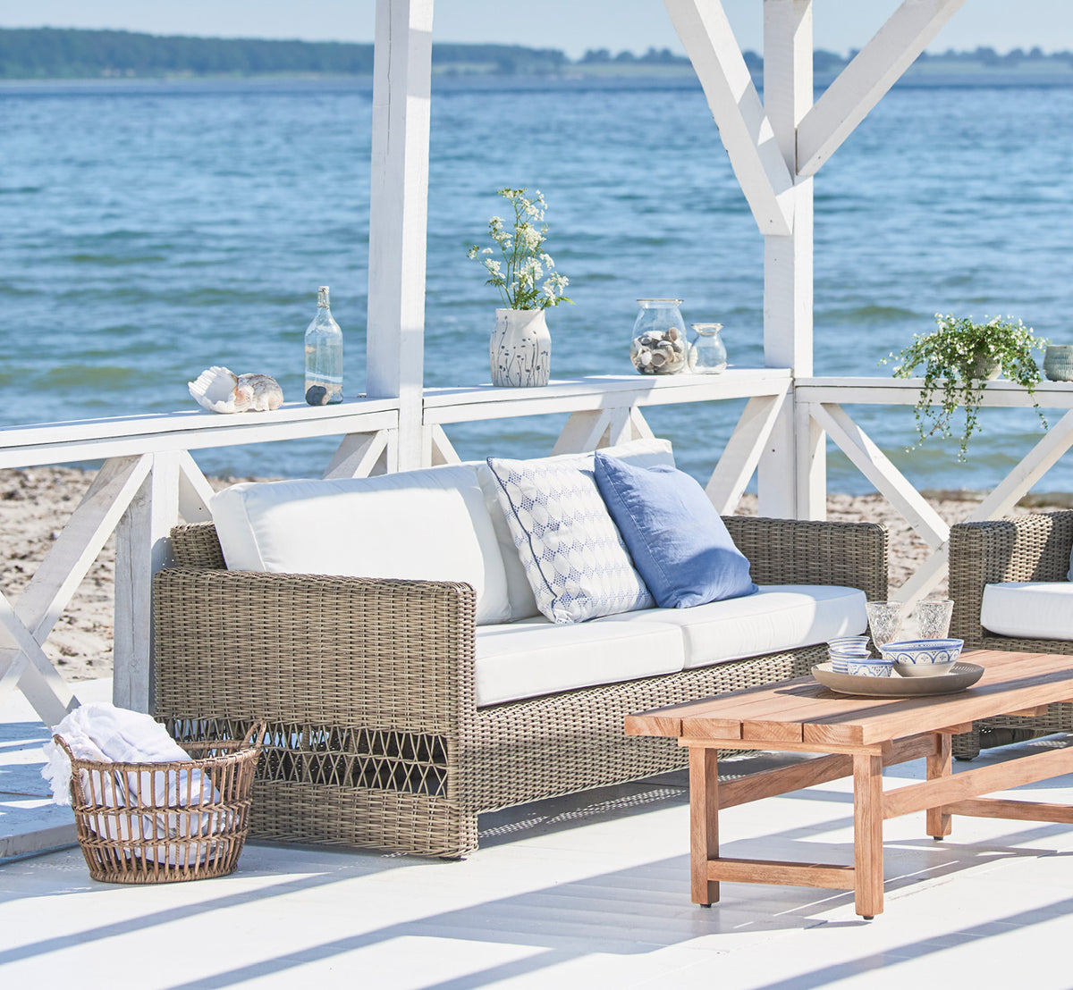 Dette er et livsstilsbillede af Carrie Sofa i antique Grey ArtFibre Aluminium, på en kystterrasse med hvidt trædæk, havudsigt og pergola.