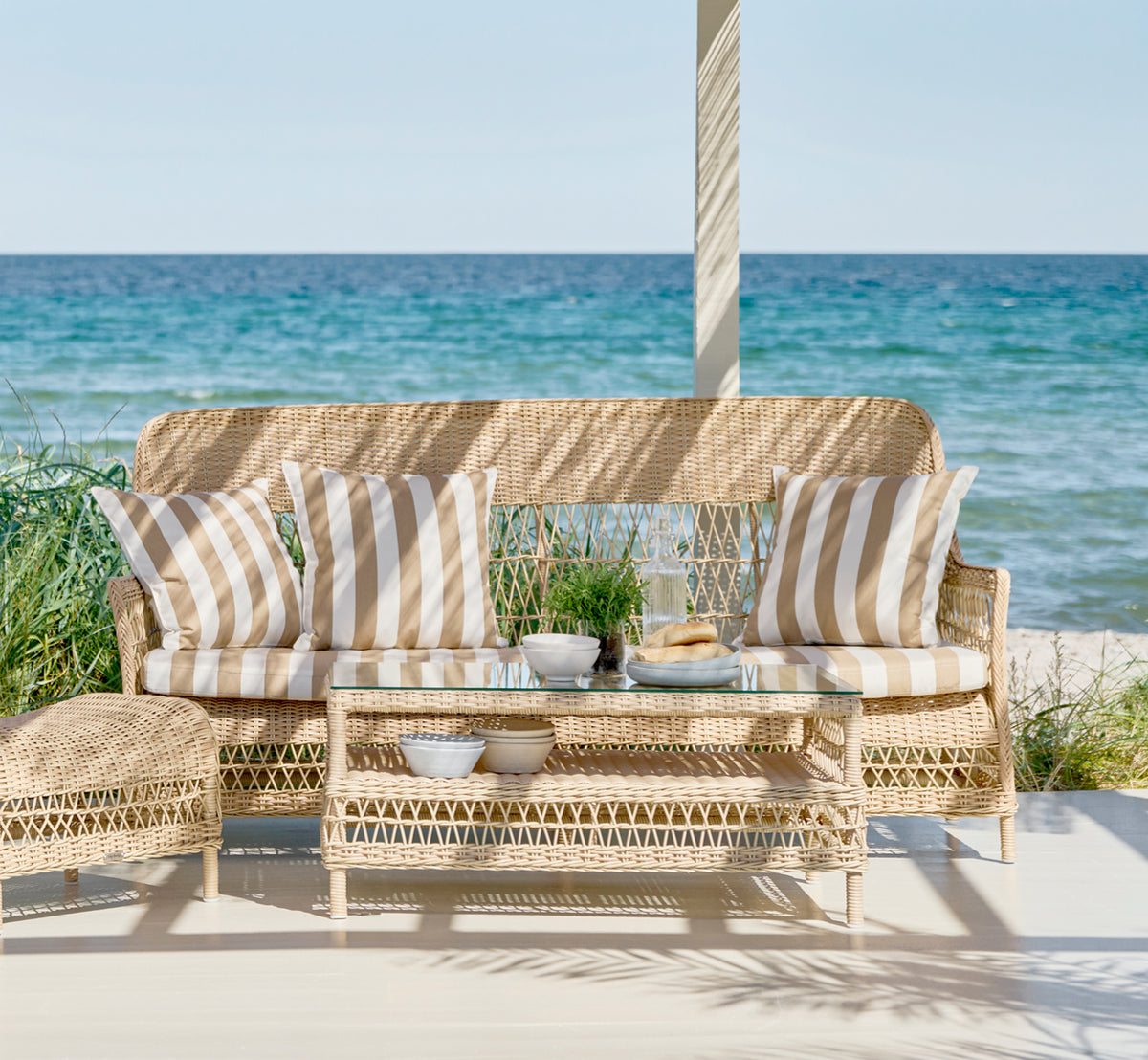 Dette er et livsstilsbillede af Dawn Sofa i natural ArtFibre Aluminium, på en kystterrasse med hvidt trædæk og havudsigt.