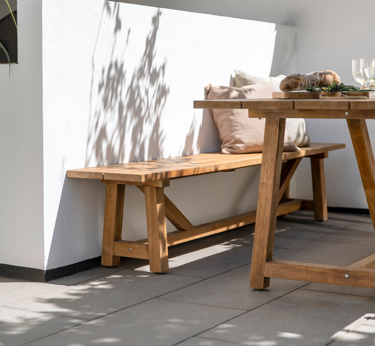 Dette er et livsstilsbillede af George Udendørs bænk i outdoor Teak, på en udendørs terrasse med grå fliser og en hvid pudset mur.