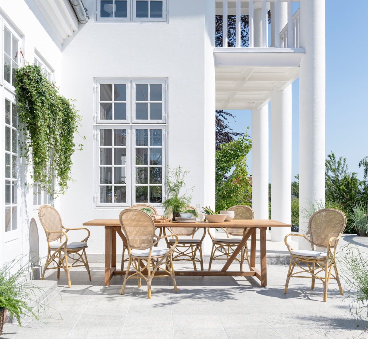 Dette er et livsstilsbillede af George Udendørs bord 240 i outdoor Teak, på en udendørs haveterrasse med hvid husfacade og grønne planter.