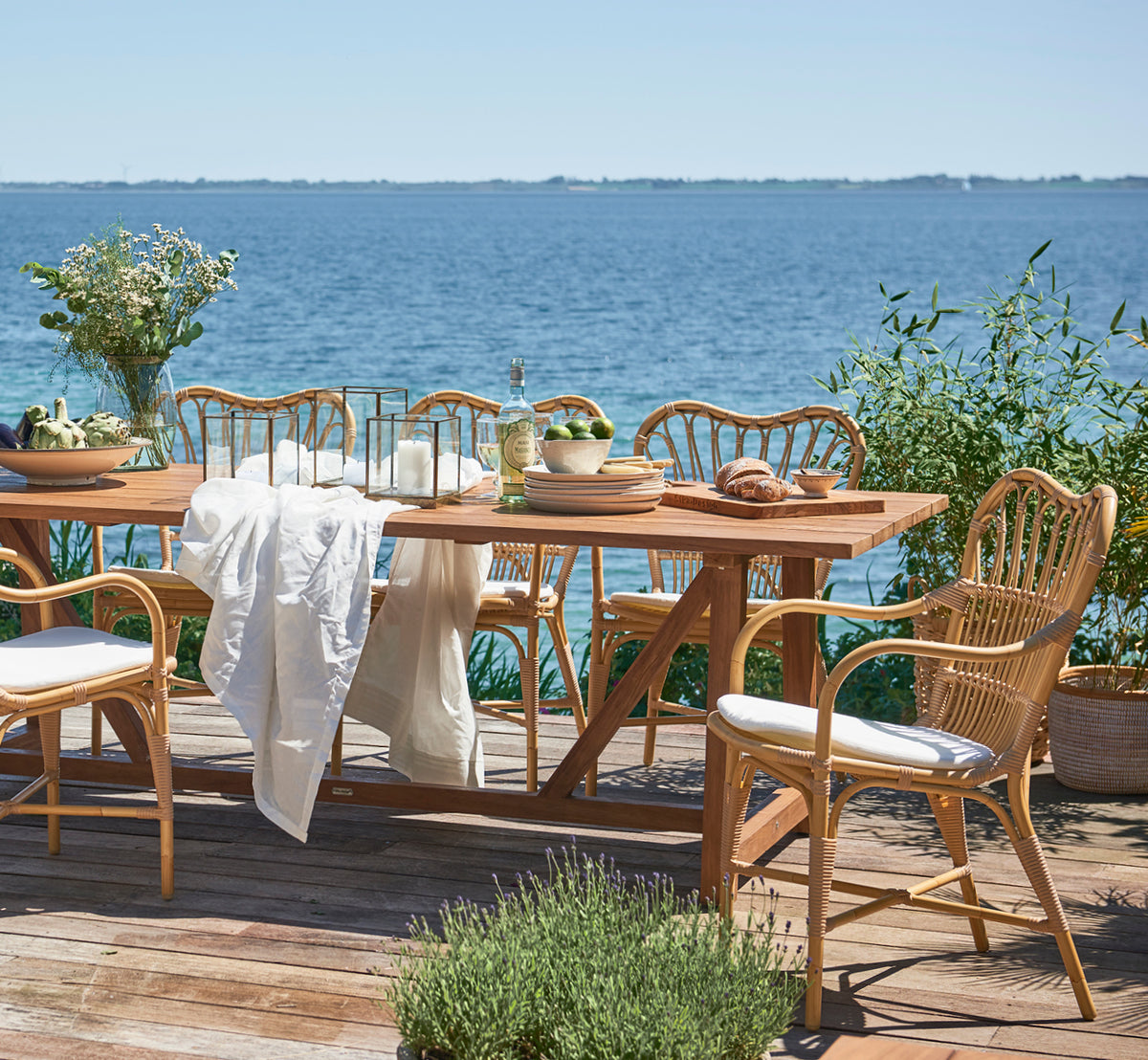 Dette er et livsstilsbillede af George Udendørs bord 240 i outdoor Teak, på en udendørs haveterrasse med hvid husfacade og grønne planter.