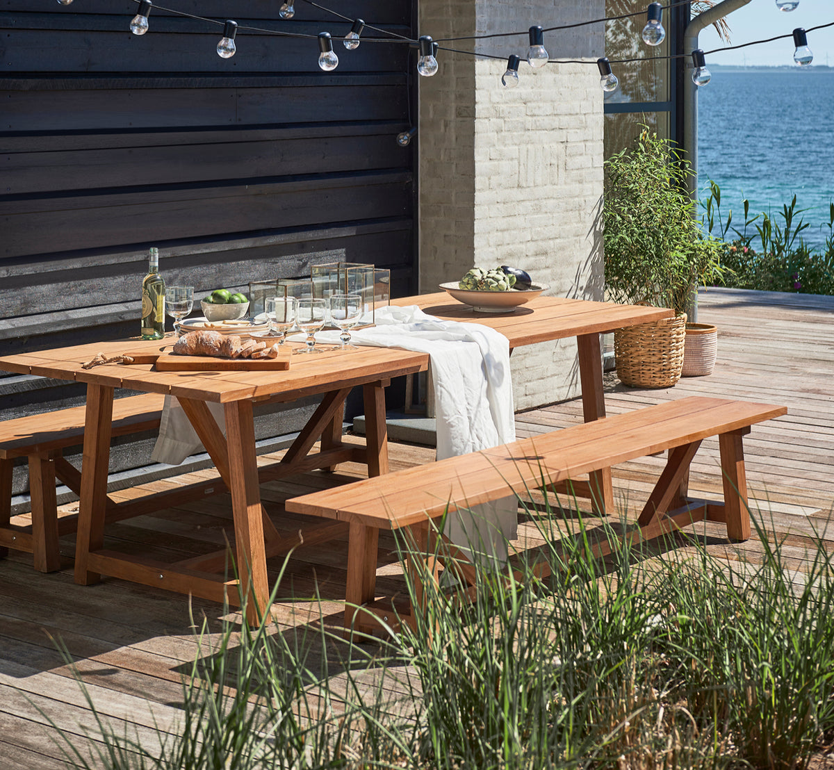 Dette er et livsstilsbillede af George Udendørs bord 240 i outdoor Teak, på en udendørs haveterrasse med hvid husfacade og grønne planter.