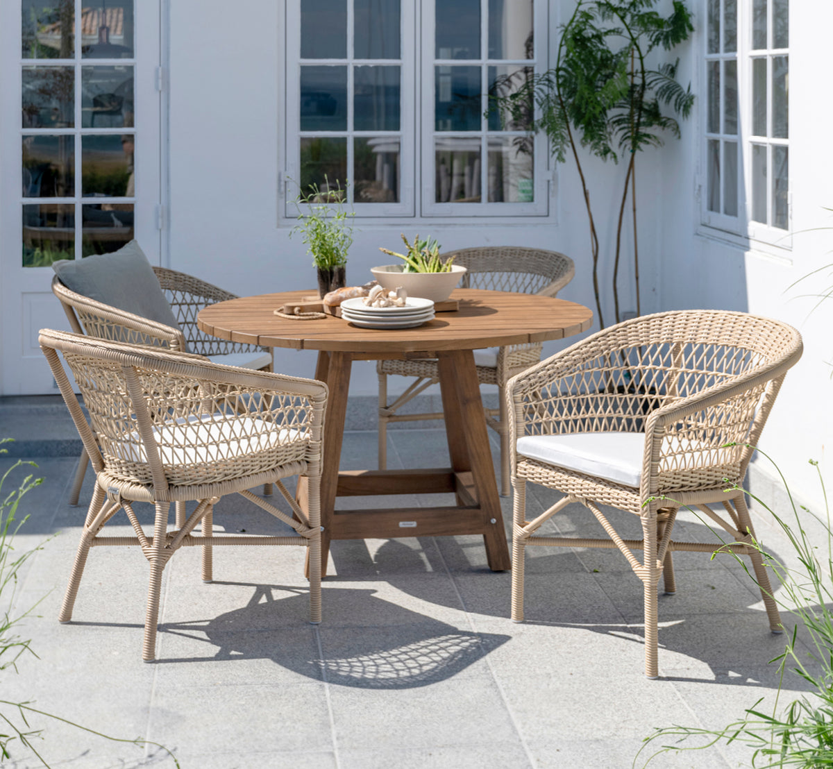 Dette er et livsstilsbillede af George Udendørs bord Ø120 i outdoor Teak, på en udendørs haveterrasse med hvid husfacade og grønne planter.