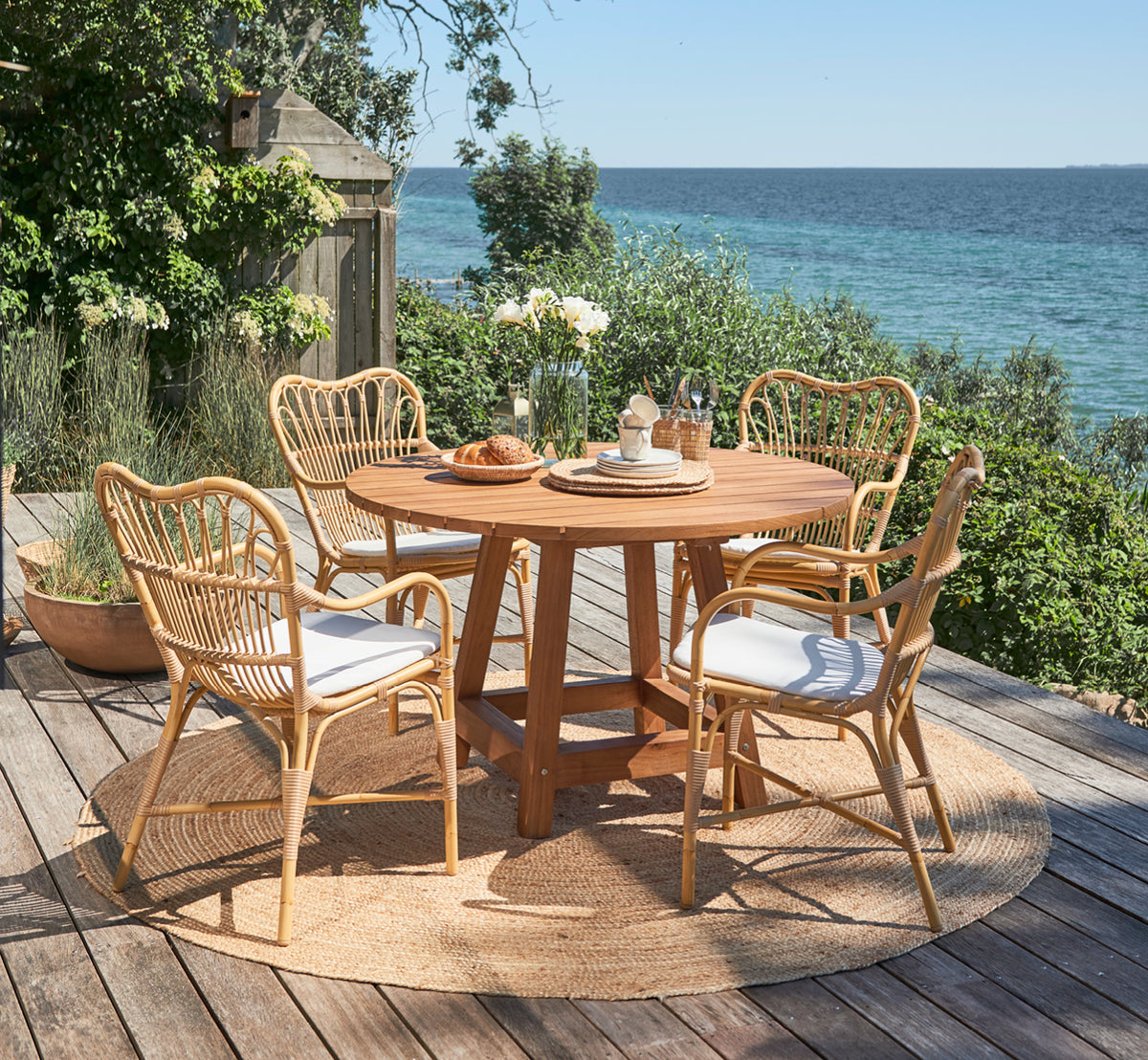 Dette er et livsstilsbillede af George Udendørs bord Ø120 i outdoor Teak, på en udendørs haveterrasse med hvid husfacade og grønne planter.