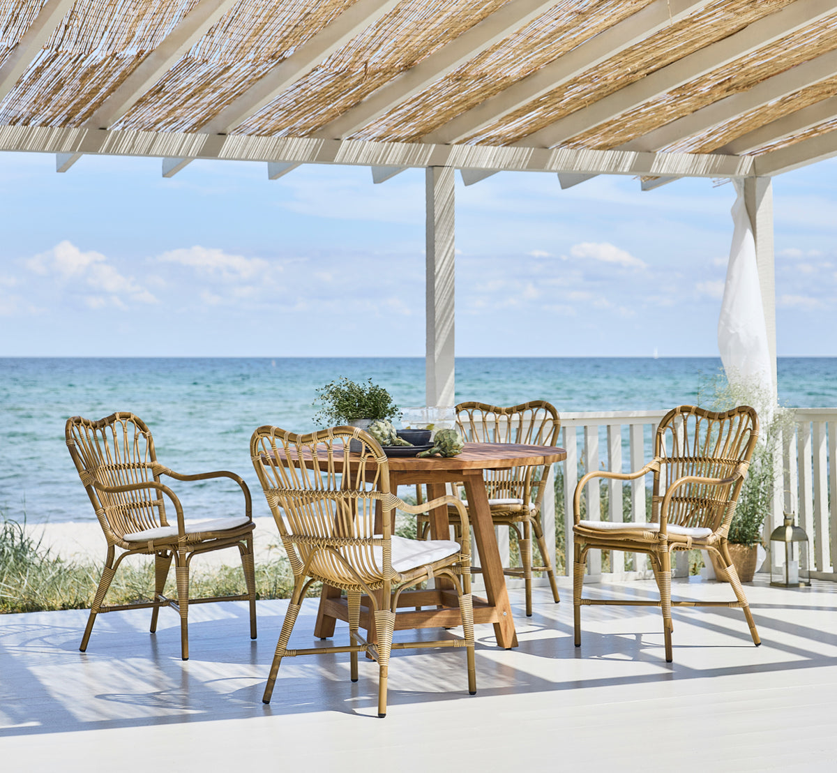 Dette er et livsstilsbillede af George Udendørs bord Ø120 i outdoor Teak, på en udendørs haveterrasse med hvid husfacade og grønne planter.