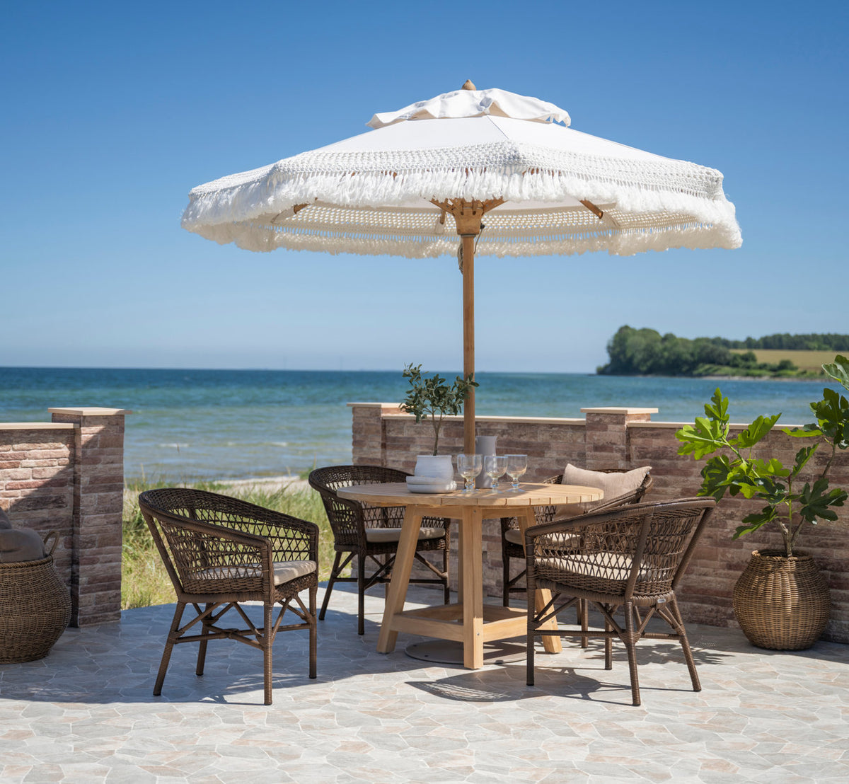 Dette er et livsstilsbillede af George Teakbord Ø120 cm i teak outdoor. Spisegruppe under en parasol med frynser ved havet.
