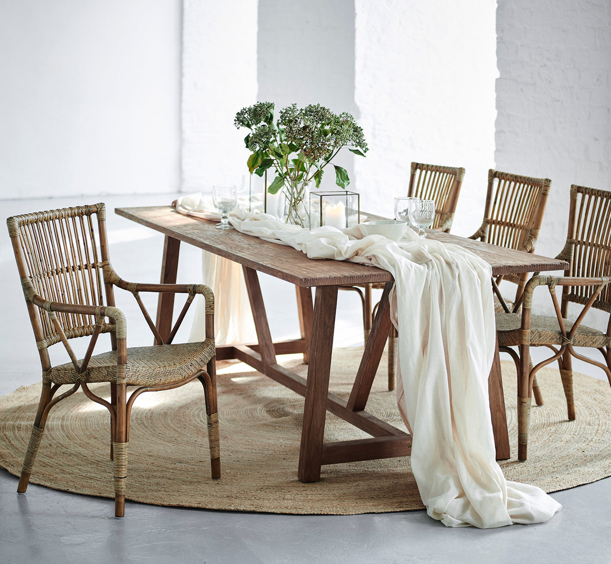 Dette er et livsstilsbillede af Lucas Teak Dining Table 240X100 cm i teak indoor. Draperet med linned og grønt, omgivet af rattanstole.