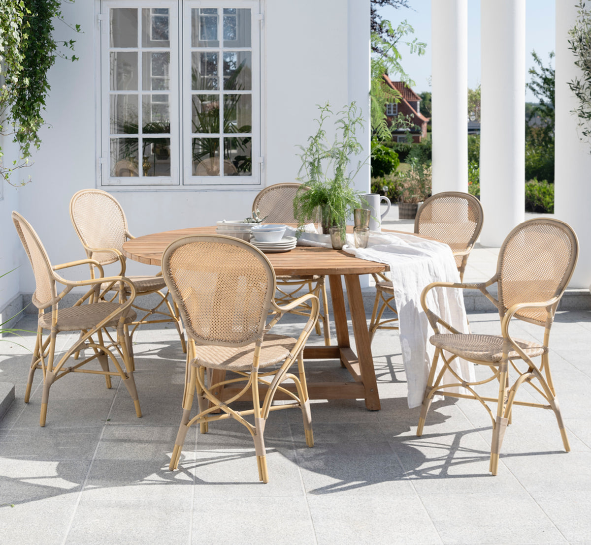 Dette er et livsstilsbillede af George Udendørs bord Ø160 i outdoor Teak, på en udendørs haveterrasse med hvid husfacade og grønne planter.