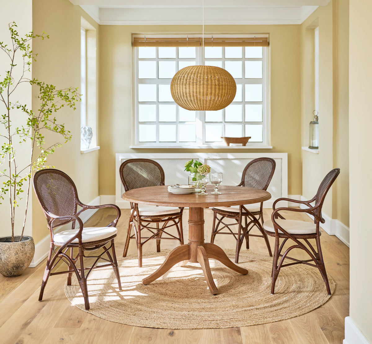 Dette er et livsstilsbillede af Michel Teak spisebord Ø120 cm i teak indoor. Med rattanstole og pendel i en solrig spisestue.