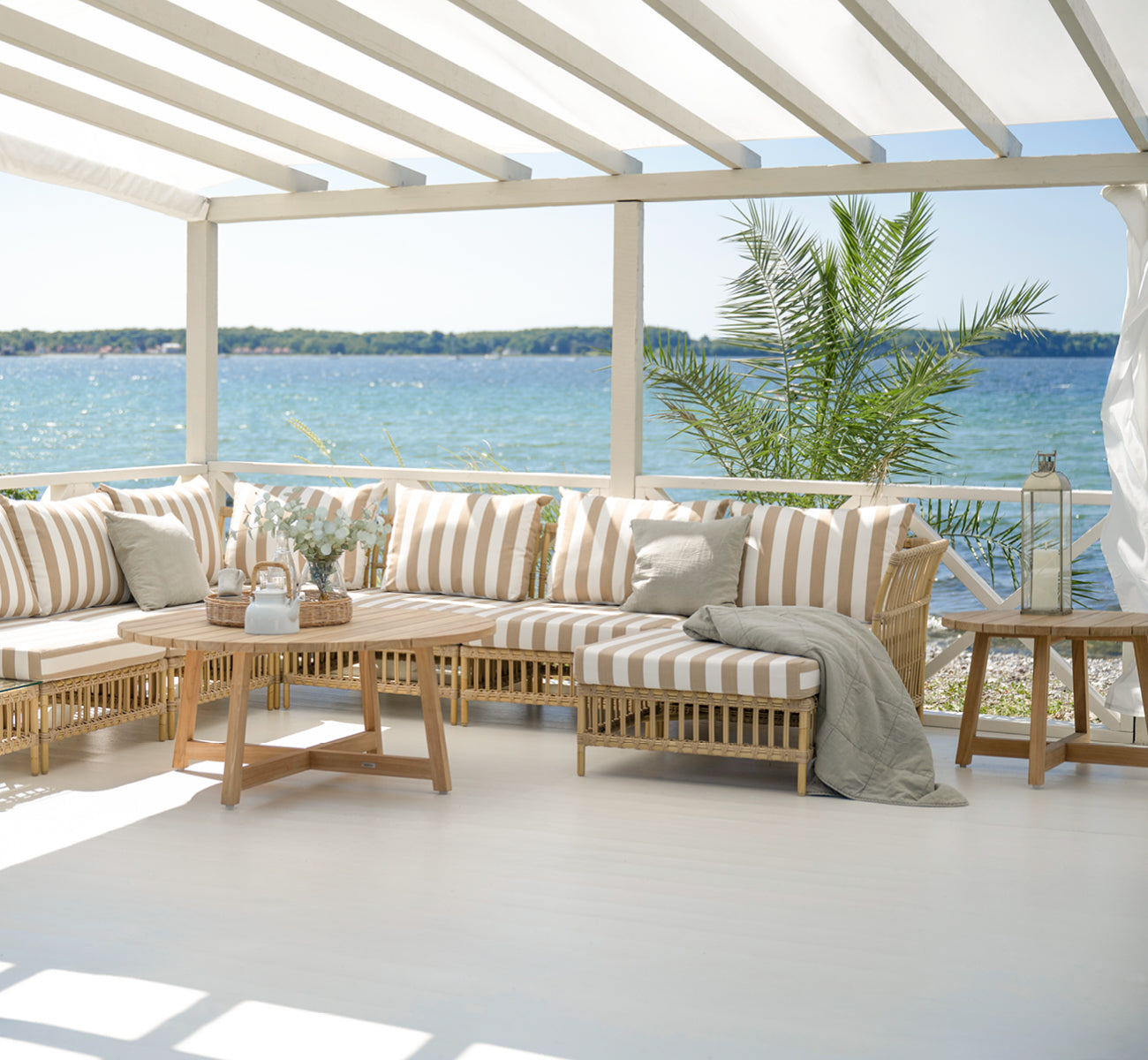 Dette er et livsstilsbillede af Victor Teak Lounge Table Ø75 cm i teak outdoor. Opstillet på en strandterrasse mellem daybeds med stribede puder.