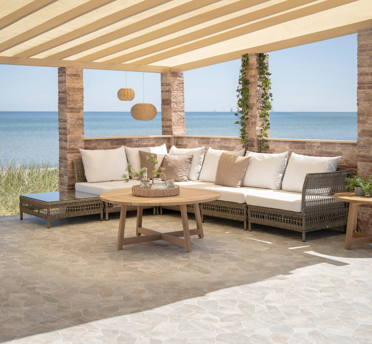 Dette er et livsstilsbillede af Victor Teak loungebord Ø75 cm i teak outdoor. Hjørnesofa med teakborde under en pergola ved havet.