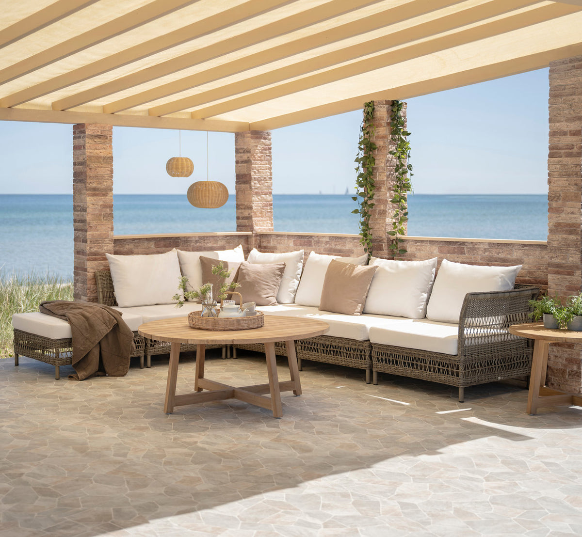 Dette er et livsstilsbillede af Victor Teak loungebord Ø75 cm i teak outdoor. Hjørnesofa, puf og teakbord under en pergola ved havet.