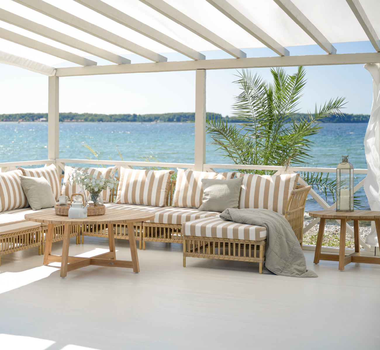 Dette er et livsstilsbillede af Victor Teak Lounge Table Ø120 cm i teak outdoor. Placeret mellem udendørs sofaer med stribede puder med udsigt over havet.