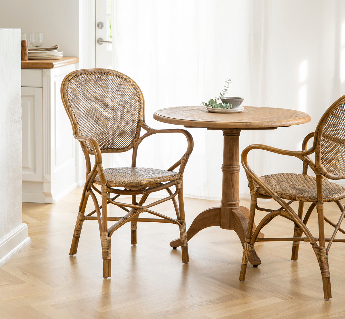 Dette er et livsstilsbillede af Michel Teak Dining Table Ø80 cm i teak indoor. Kombineret med rattanlænestole i et lyst og luftigt caféinteriør.
