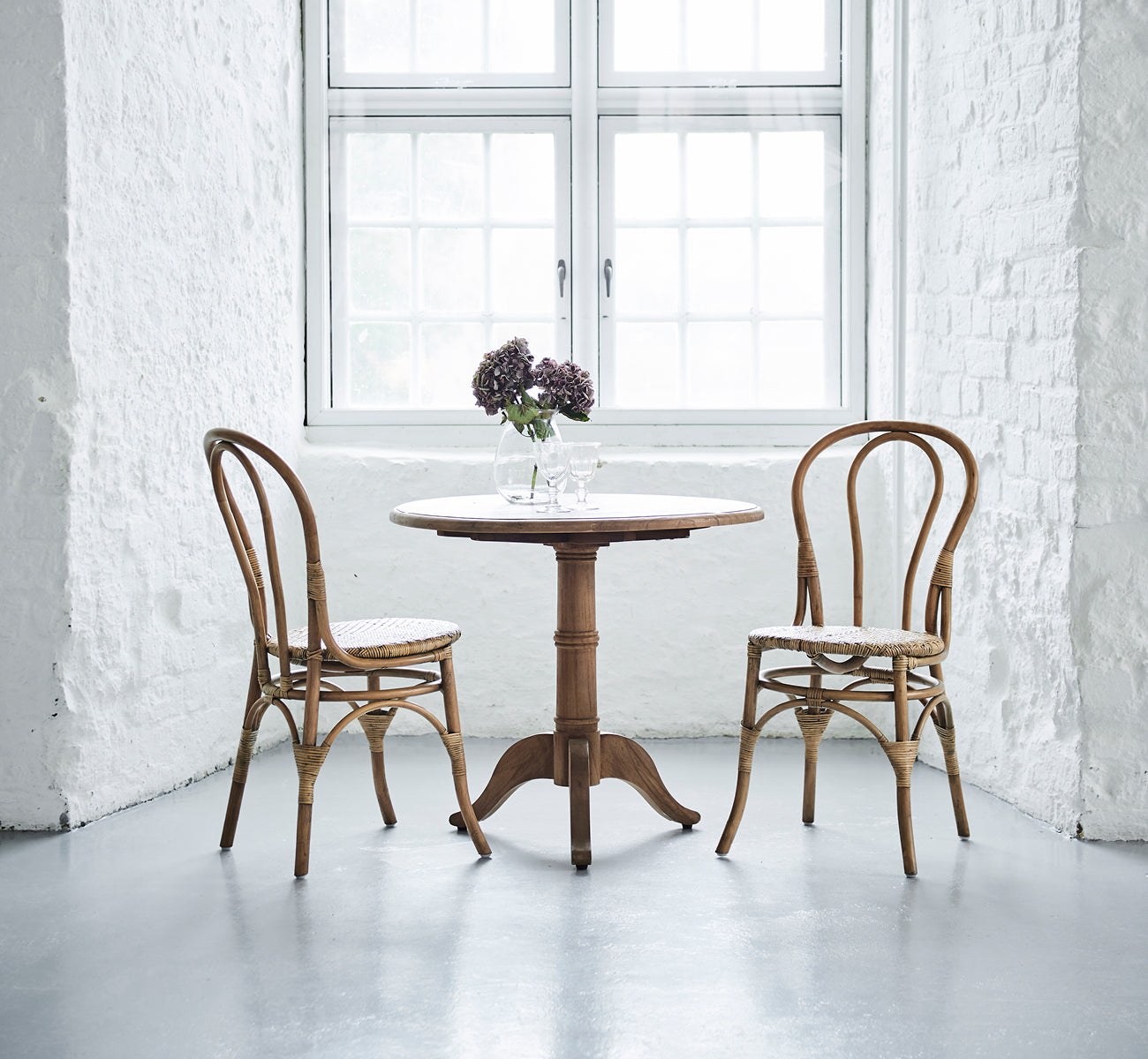 Dette er et livsstilsbillede af Michel Teak Dining Table Ø80 cm i teak indoor. Dækket med to stole og blomster i et lyst hvidt murstensinteriør.
