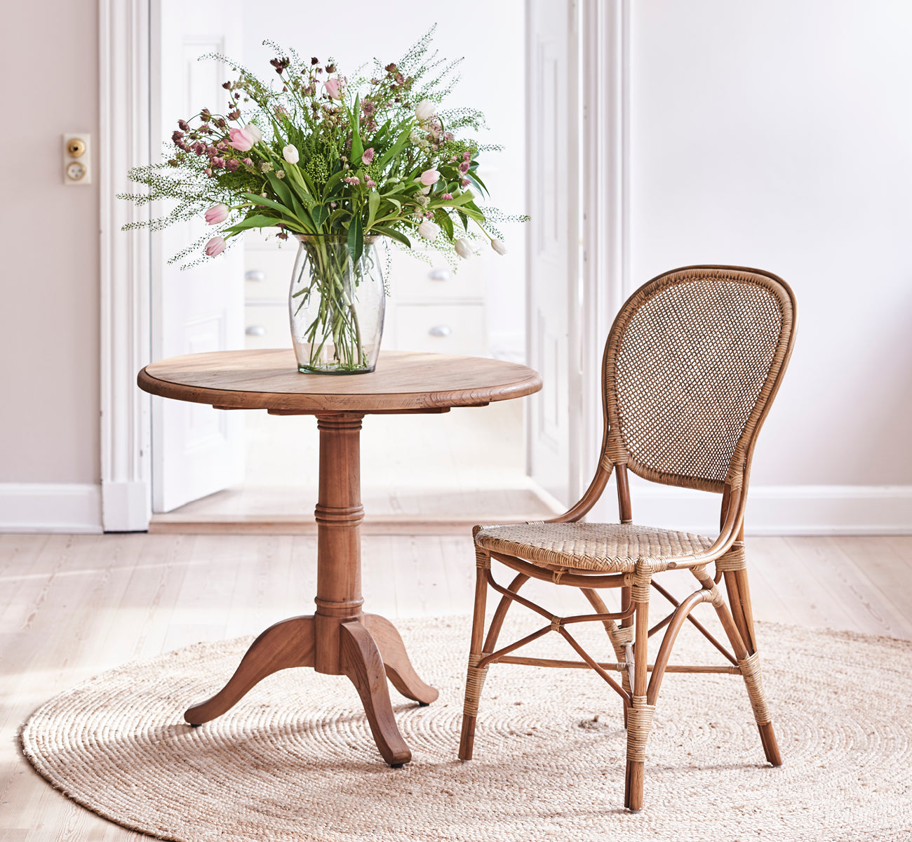 Dette er et livsstilsbillede af Michel Teak Dining Table Ø80 cm i teak indoor. Opstillet i et varmt rustikt spisemiljø med rattanstole og blødt lys.