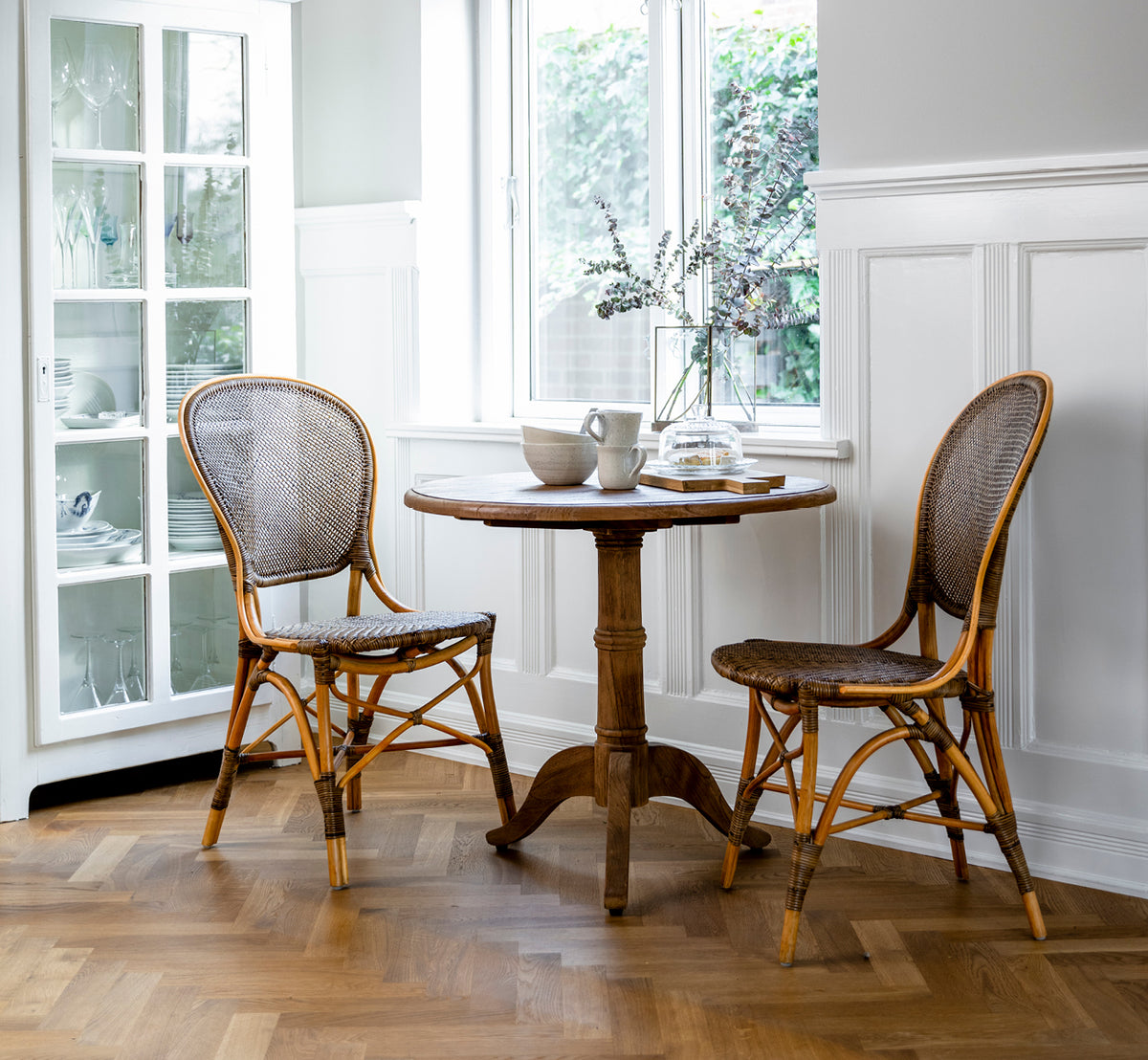 Dette er et livsstilsbillede af Michel Teak Dining Table Ø80 cm i teak indoor. Opstillet i et varmt rustikt spisemiljø med rattanstole og blødt lys.