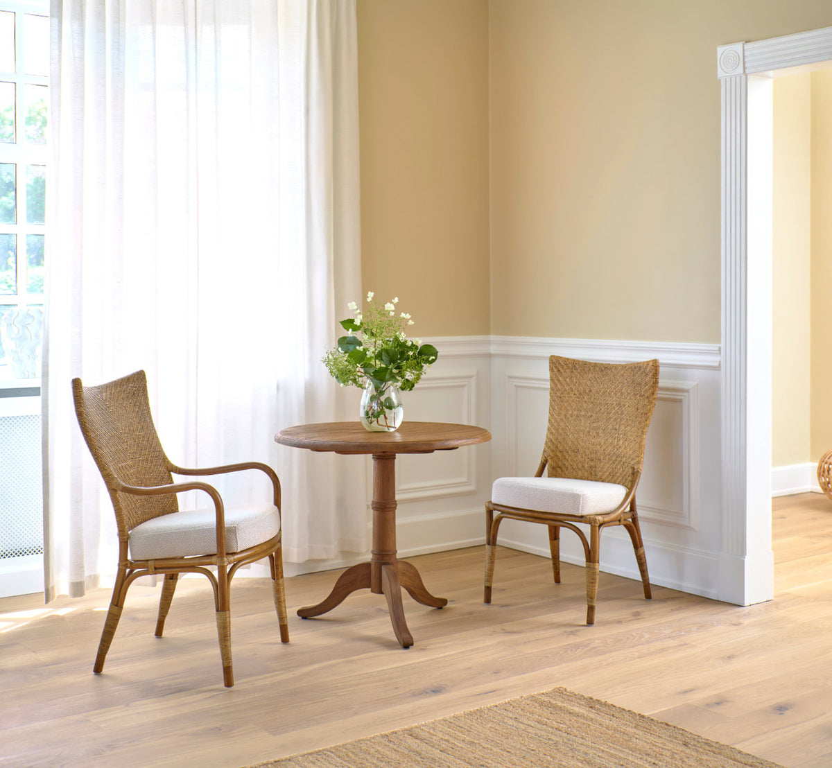 Dette er et livsstilsbillede af Michel Teak spisebord Ø80 cm i teak indoor. Med rattanstole ved hvide paneler og gardiner.