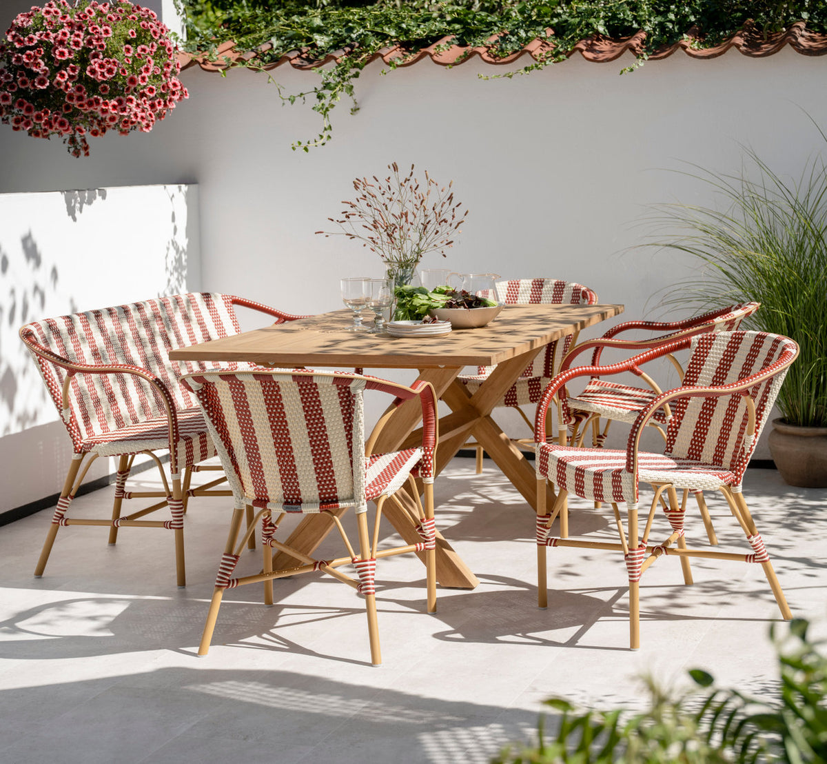Dette er et livsstilsbillede af Colonial Teakbord 160x100 cm i teak outdoor. Med stribede stole på en solrig terrasse med mur.