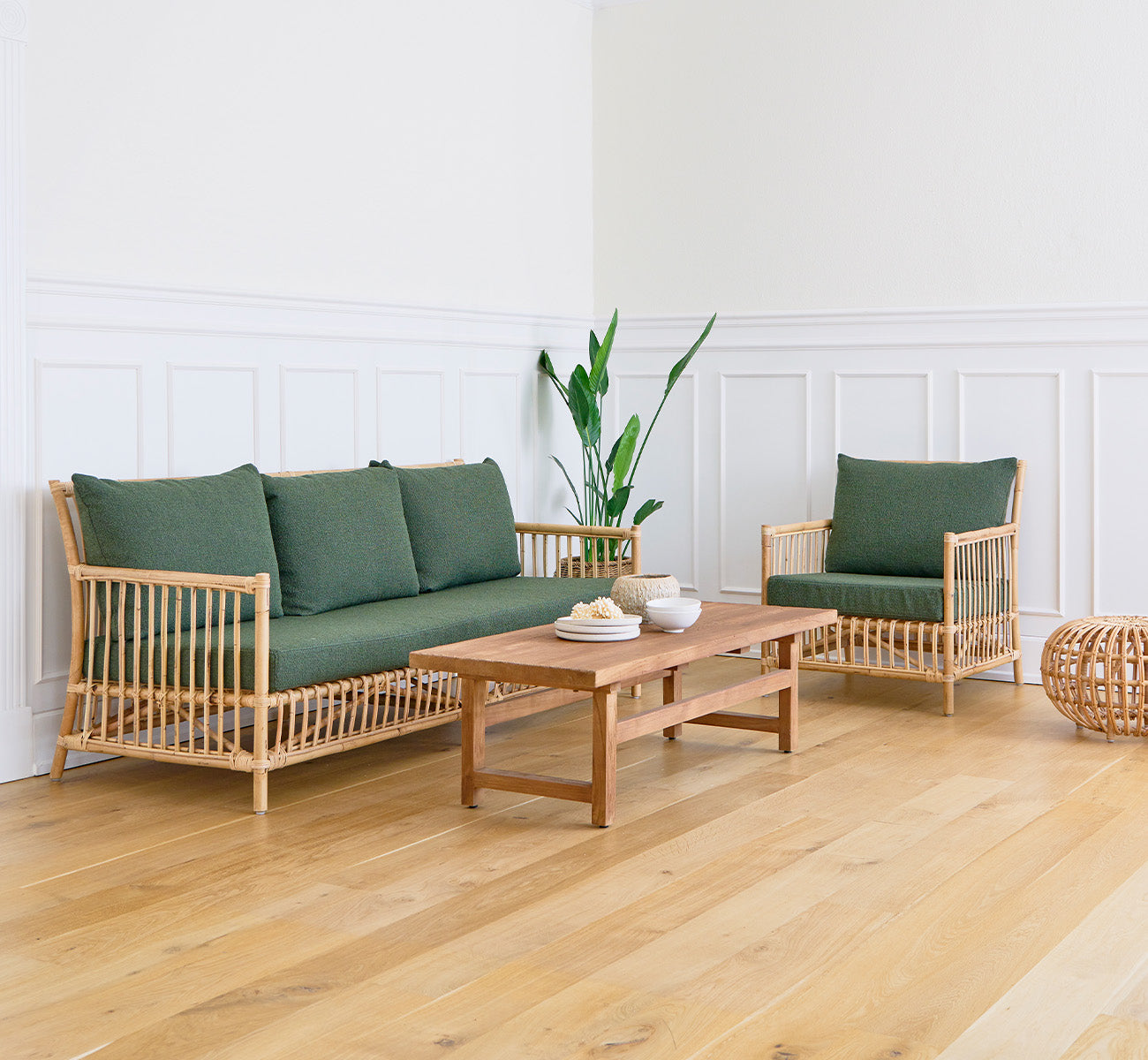 Dette er et livsstilsbillede af Alfred Sofabord i teak indoor, i en lys stue med hvide panelvægge og en sofa med grønne puder.