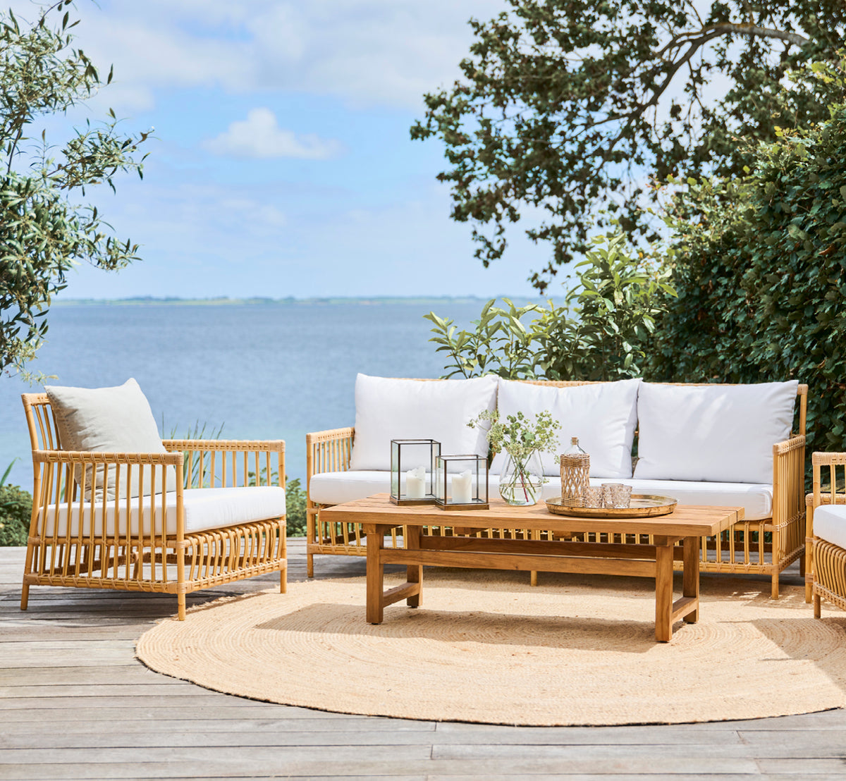 Dette er et livsstilsbillede af Julian Teak Lounge Table 140x55 cm i teak outdoor. På en terrasse med loungemøbler, et jutetæppe og havudsigt.