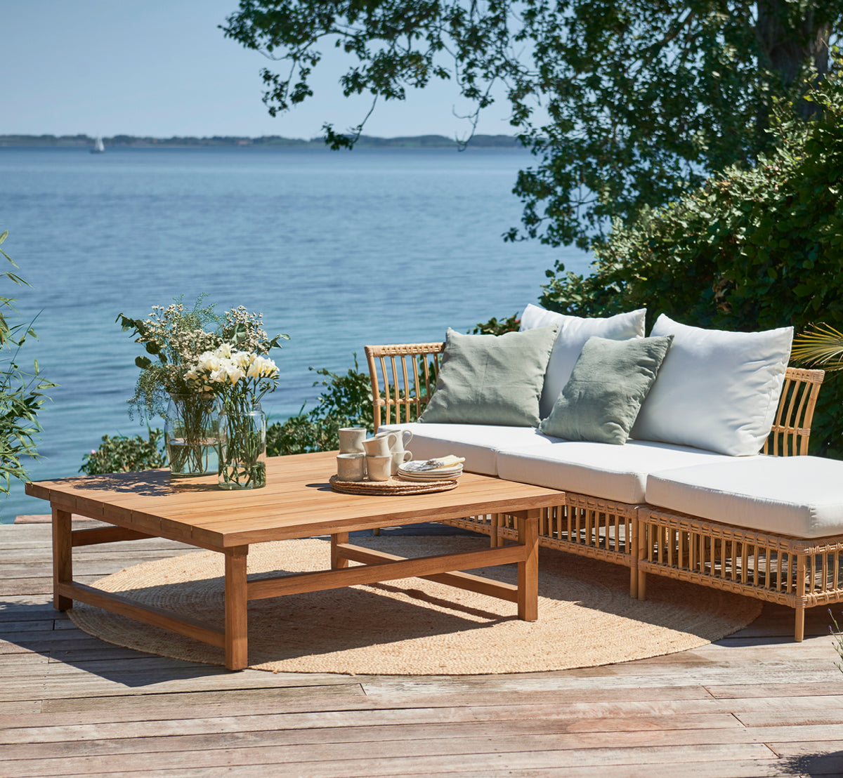 Dette er et livsstilsbillede af Julian Teak Lounge Table 120x120 cm i teak outdoor. På en strandterrasse med en loungesofa, puder og en teservering.