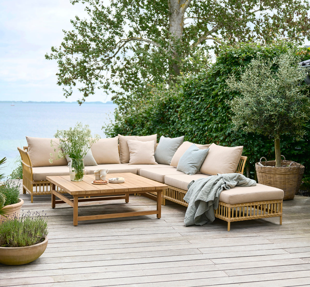 Dette er et livsstilsbillede af Julian Teak Lounge Table 120x120 cm i teak outdoor. På en haveterrasse ved havet med loungesæder og frodig beplantning.