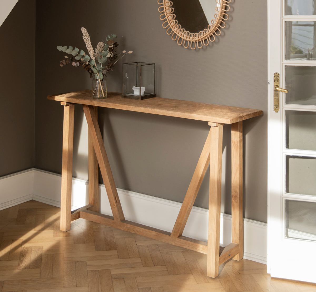 Dette er et livsstilsbillede af Lucas Teak Konsol i teak indoor. Mod en mørkegrå væg med et rattanspejl over og eukalyptus i en vase.