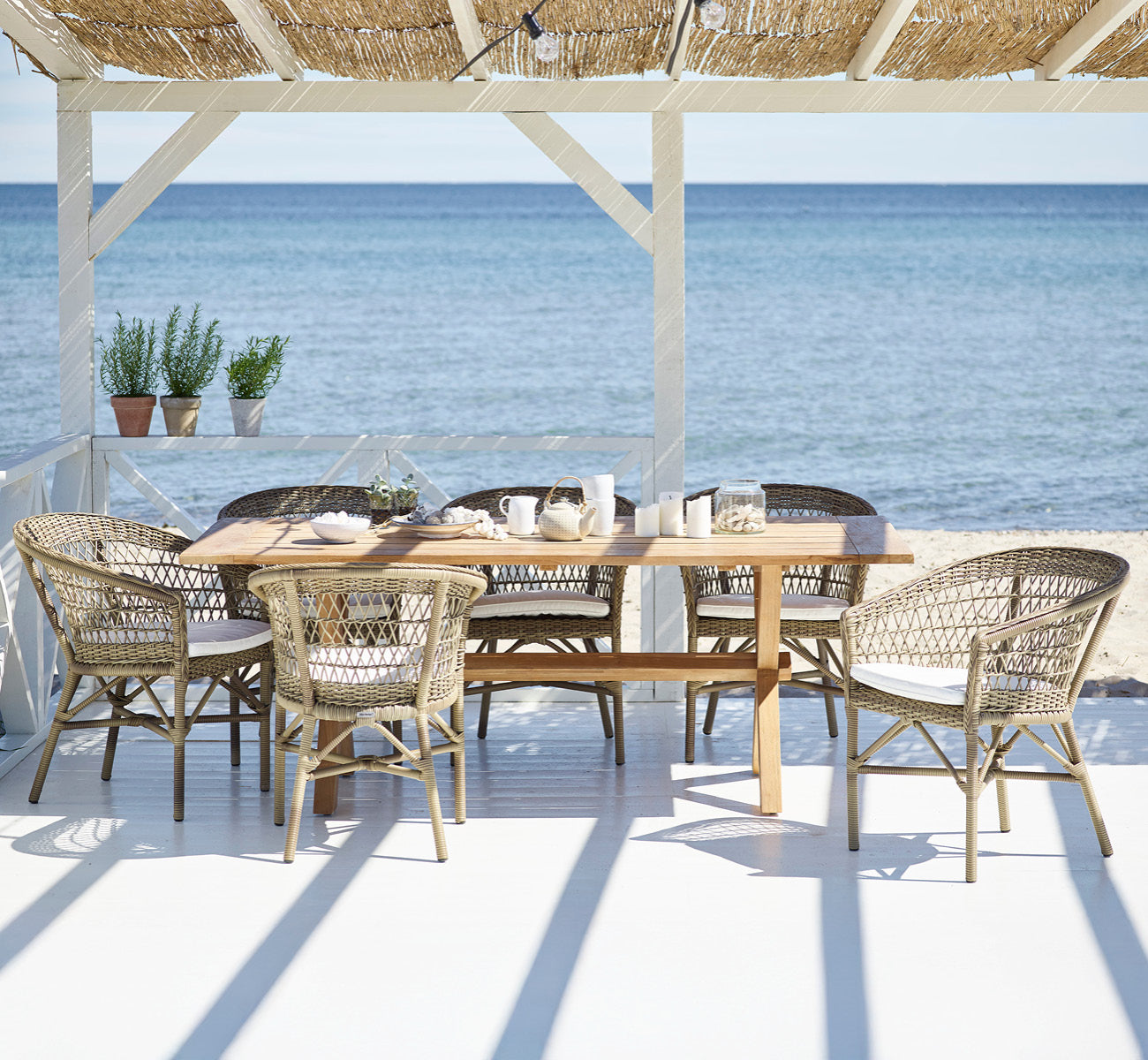 Dette er et livsstilsbillede af Colonial Bord 200 i outdoor Teak, på en strandterrasse med hvidt trædæk og havudsigt.