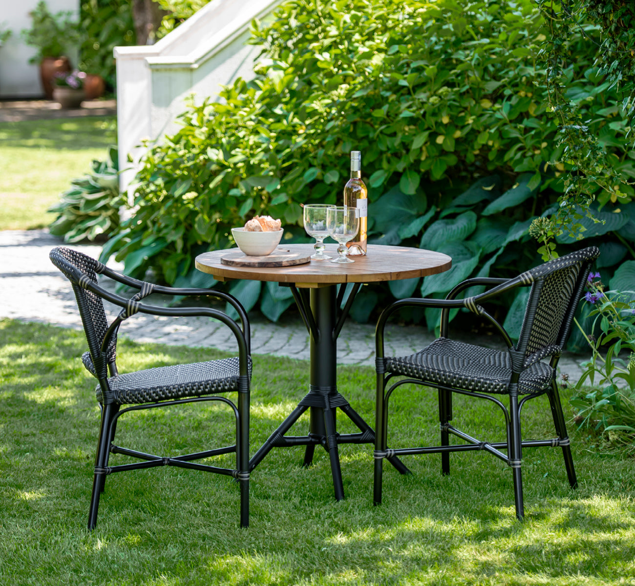 Dette er et livsstilsbillede af Nicole Cafe Table i black aluminium frame black ArtFibre. På en stenterrasse med havevegetation.
