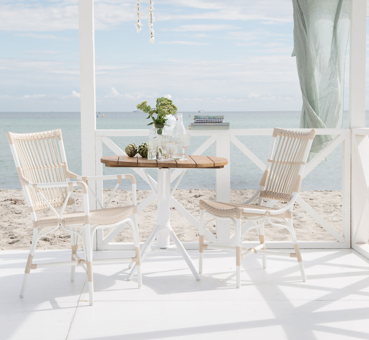 Dette er et livsstilsbillede af Nicole Cafe Table i white aluminium frame white ArtFibre. Med Piano-stole på et dæk ved havet.