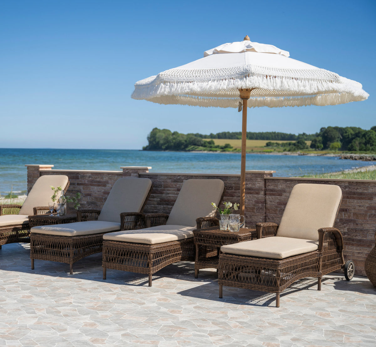 Dette er et livsstilsbillede af Susy Udendørs sidebord i brown artfibre aluminum. Række af liggestole under en parasol med frynser ved havet.