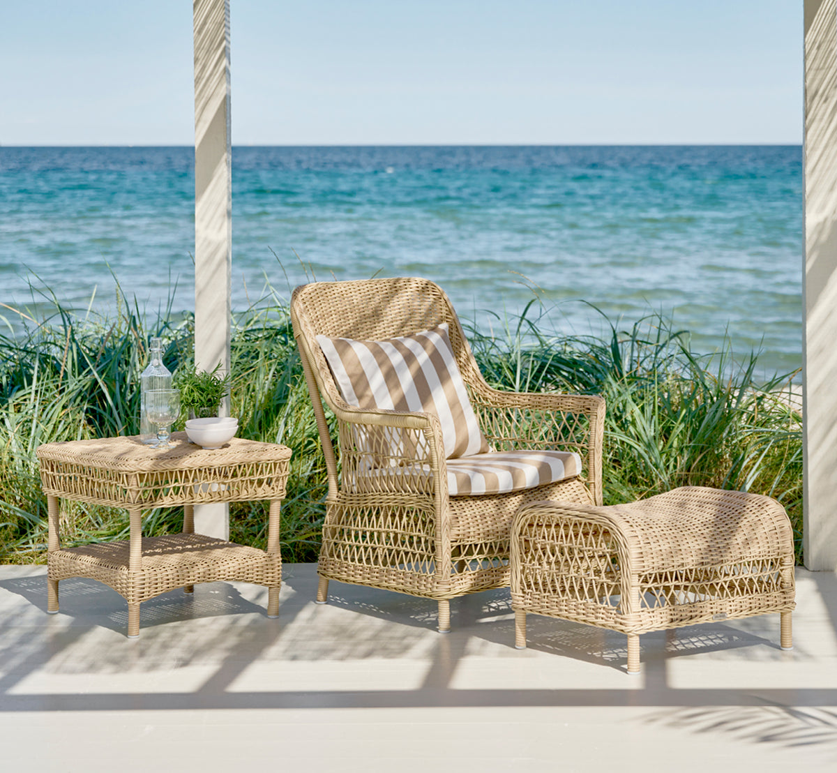 Dette er et livsstilsbillede af Susy Sidebord i natural ArtFibre Aluminium, på en kystterrasse med havudsigt, strandgræs og hvide søjler.