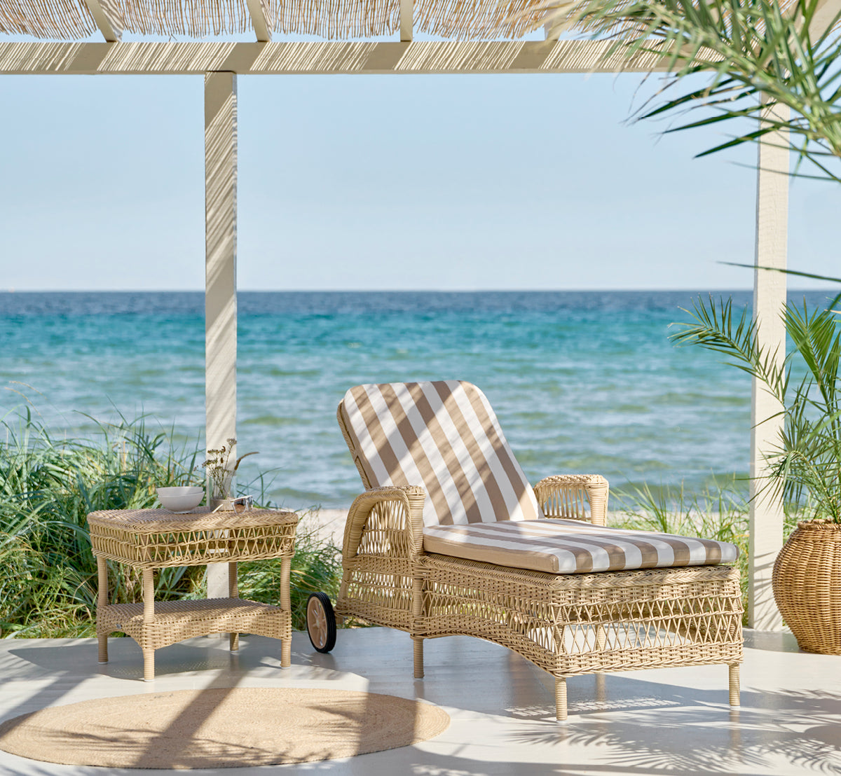 Dette er et livsstilsbillede af Susy Sidebord i natural ArtFibre Aluminium, på en kystterrasse med havudsigt, strandgræs og hvide søjler.
