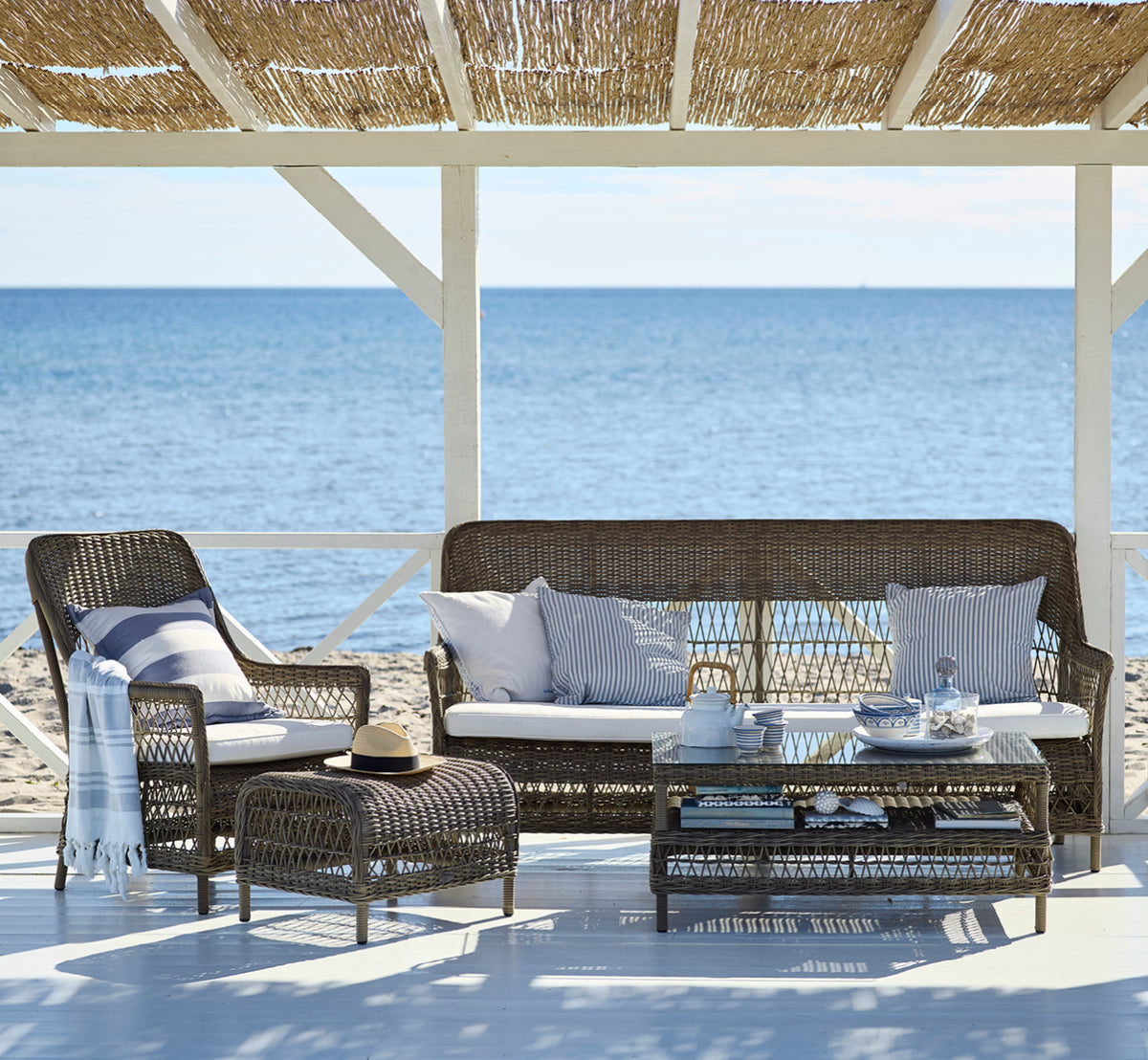Dette er et livsstilsbillede af Hazel Sofabord i antique Grey ArtFibre Aluminium, på en skygget kystterrasse med sivtag og havudsigt.