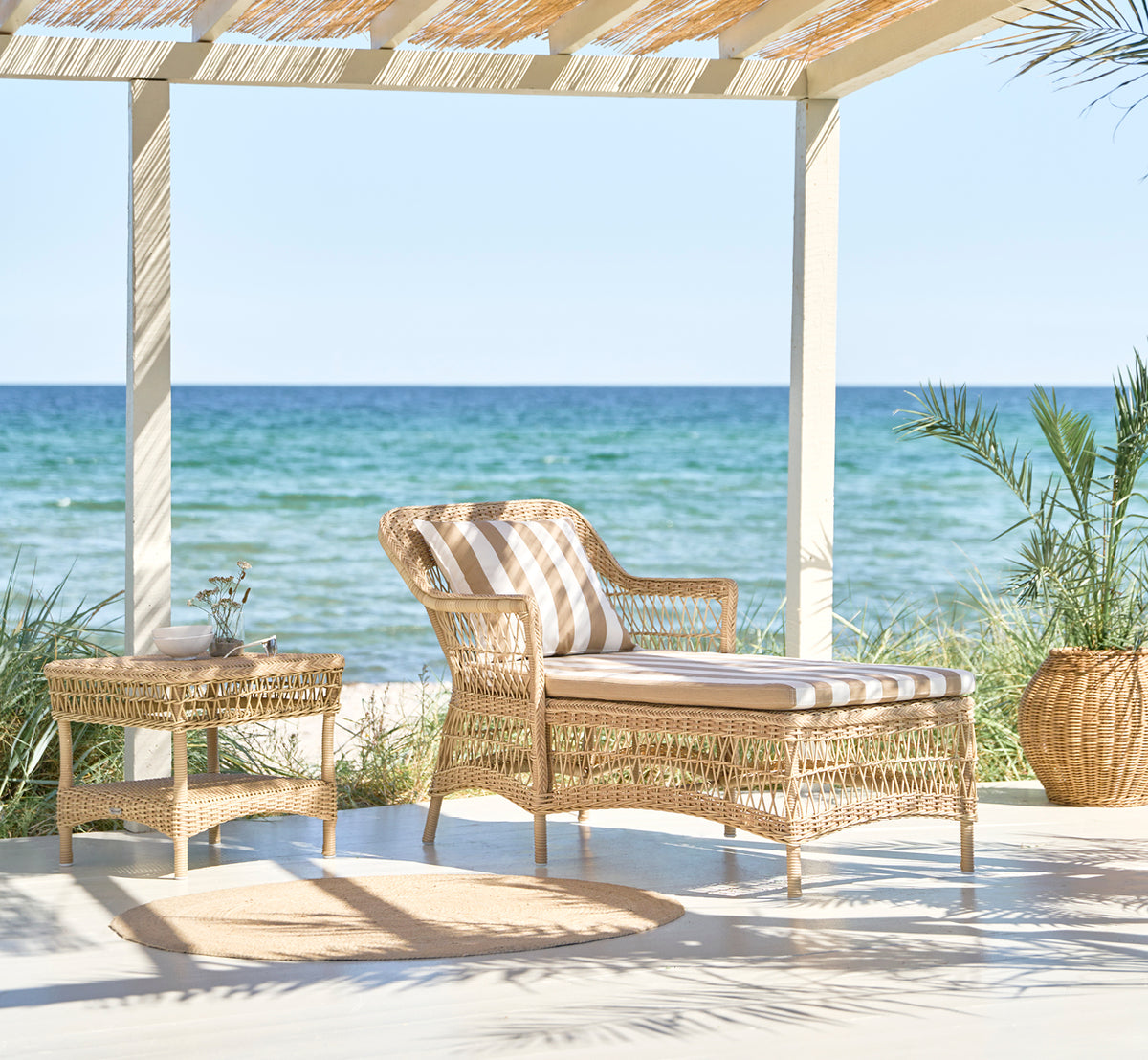 Dette er et livsstilsbillede af Olivia Exterior Sunbed i natural artfibre aluminium. I en frodig have med pude og plaid.