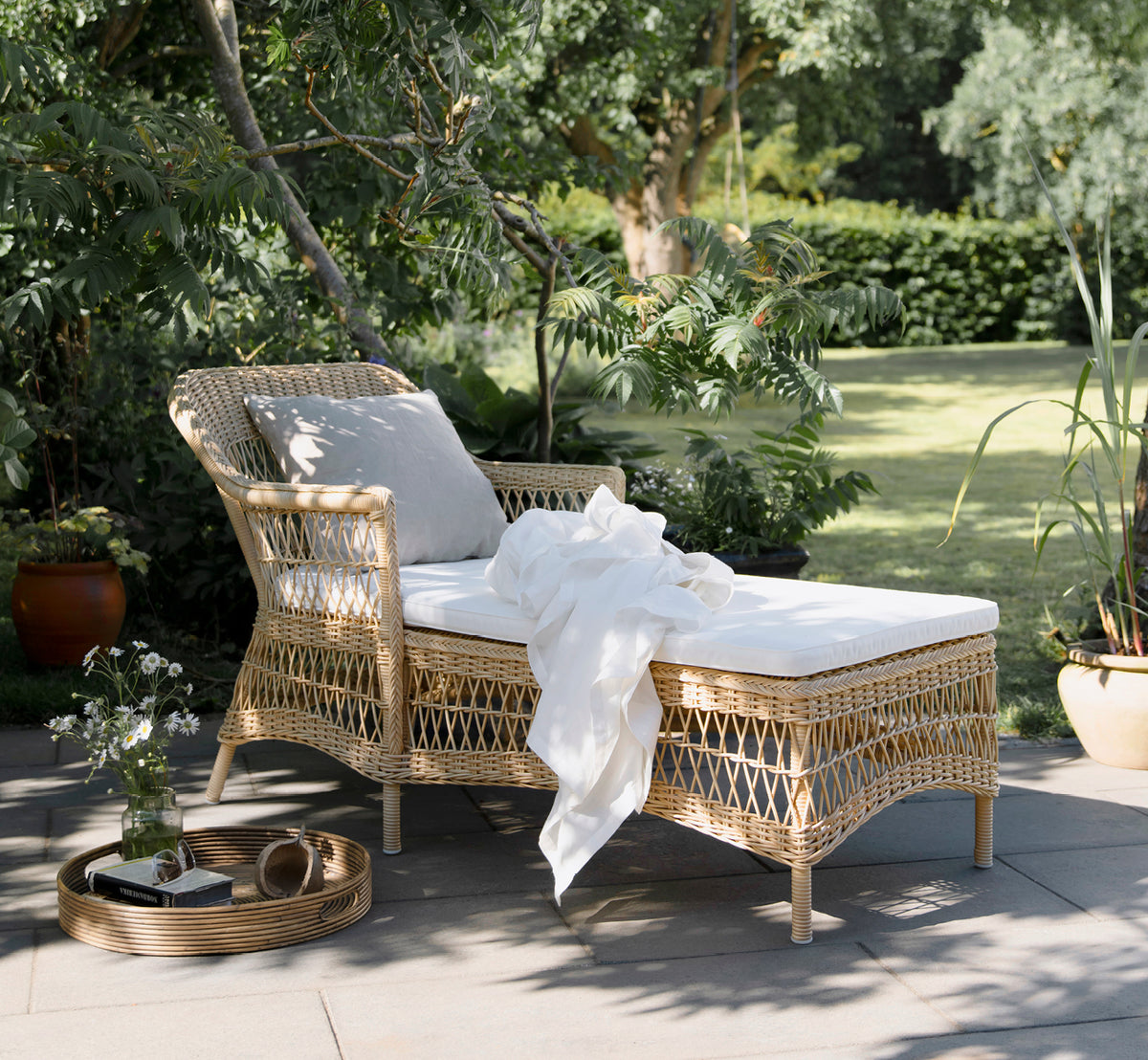 Dette er et livsstilsbillede af Olivia Exterior Sunbed i natural artfibre aluminium. I en havescene med hvidt tæppe og pude.