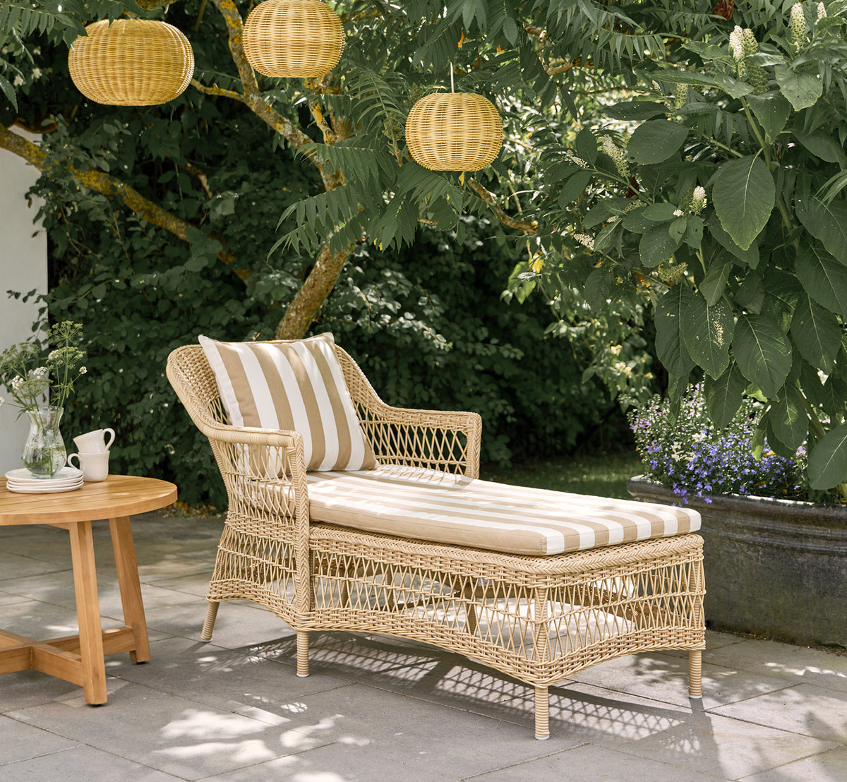 Dette er et livsstilsbillede af Olivia Exterior Sunbed i natural artfibre aluminium. På en terrasse med stribet pude og lanterner.