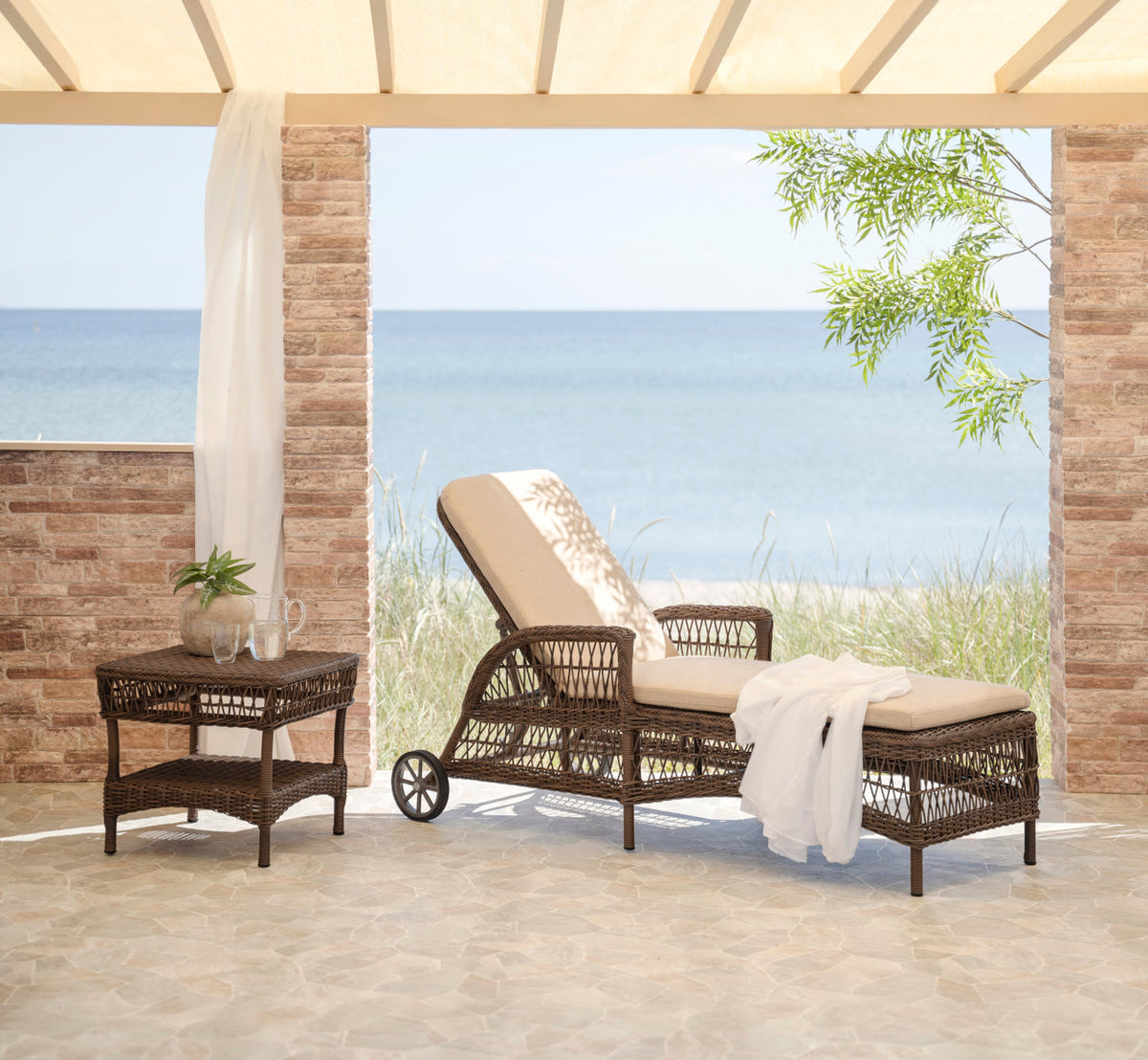 Dette er et livsstilsbillede af Daisy Udendørs liggestol i brown artfibre aluminum. Med et hvidt håndklæde på en terrasse ved havet.