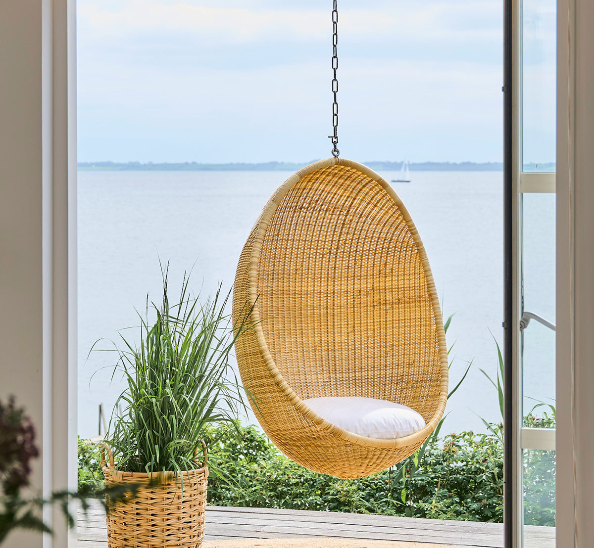 Dette er et livsstilsbillede af Chain Hængeæg i metal, hængende ved en glasdør med vandudsigt og omgivende grønne planter.