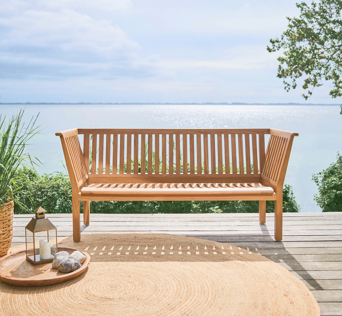 Dette er et livsstilsbillede af St Catherine Teakbænk i teak outdoor. Opstillet på et trædæk med havudsigt og et rundt jutetæppe.