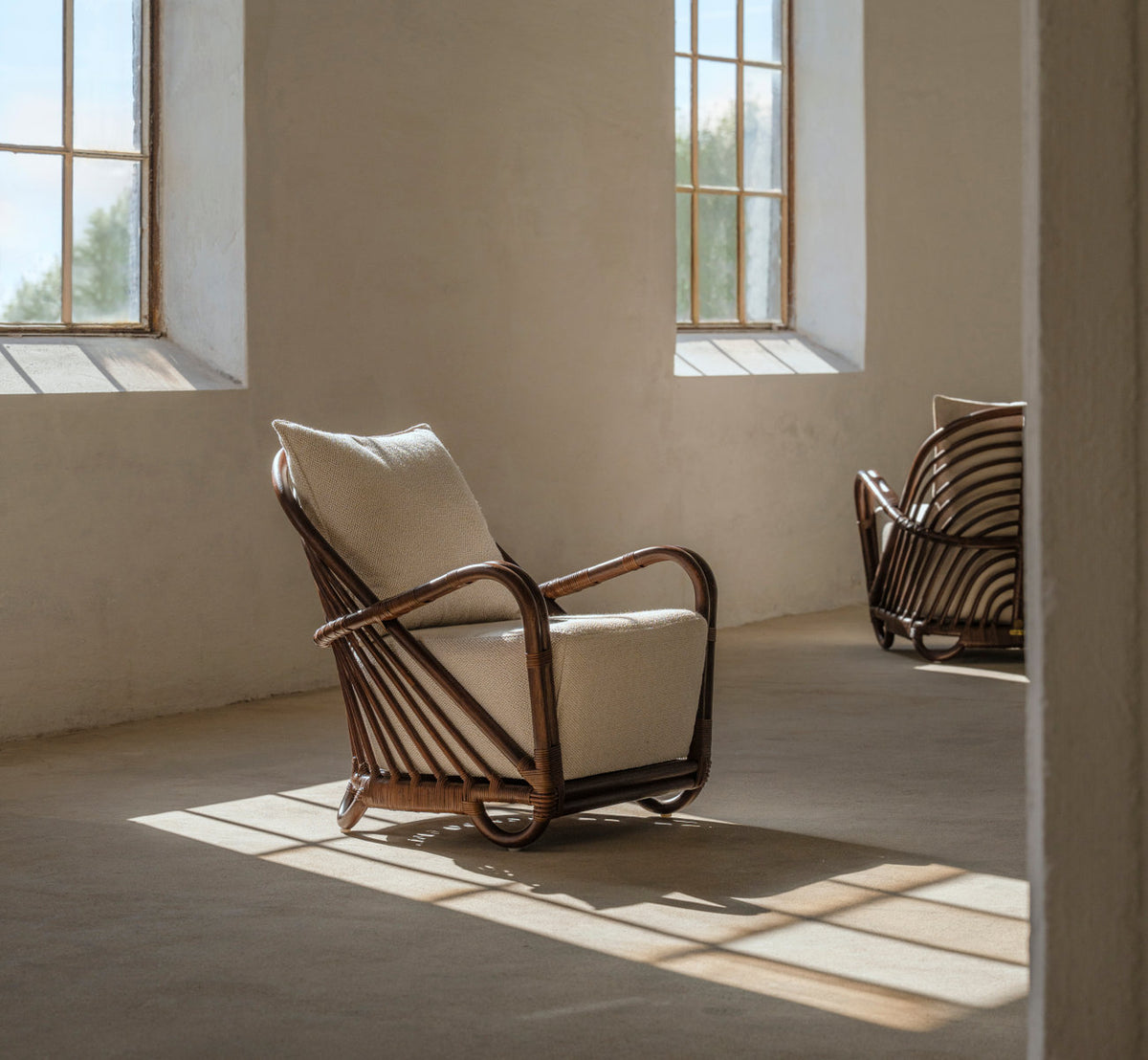 Dette er et livsstilsbillede af Charlottenborg Lænestol i brown rattan. Med puder i et solbeskinnet rum med betongulv.