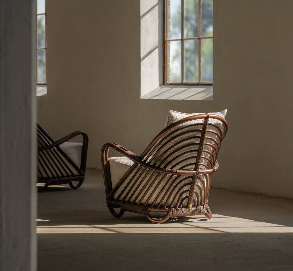 Dette er et livsstilsbillede af Charlottenborg Lænestol i brown rattan. To i blødt vindueslys med dybe skygger.