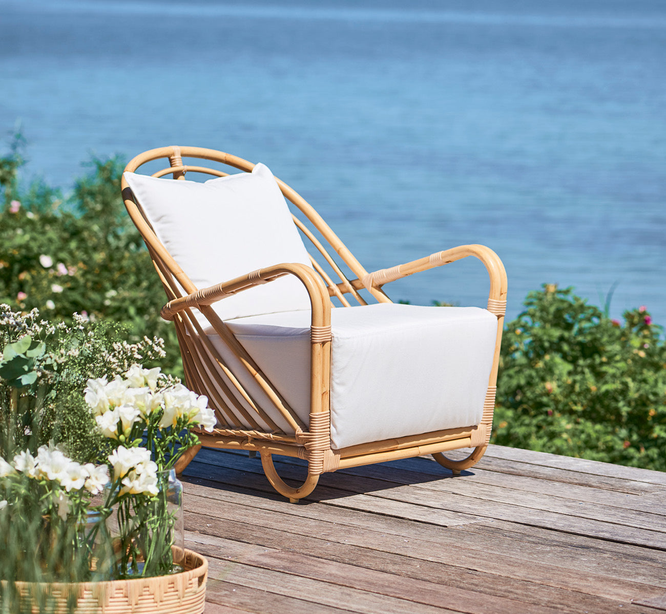 Dette er et livsstilsbillede af Charlottenborg Udendørs lænestol i natural Aluminium ArtFibre, på et udendørs trædæk med blomster og havudsigt.