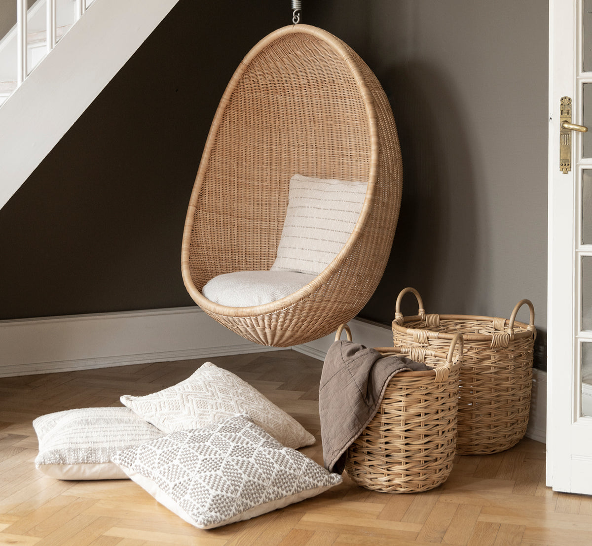 Dette er et livsstilsbillede af Sarah Kurvesæt i natural rattan. Placeret ved siden af en hængende podstol og mønstrede puder på parket.