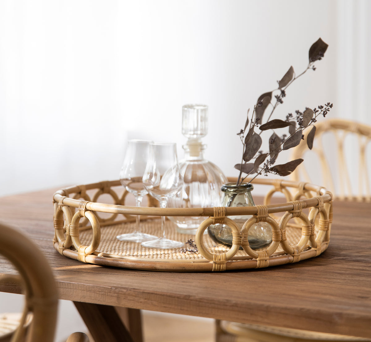 Dette er et livsstilsbillede af Mira Tray i natural rattan. På et spisebord med glasvarer og tørret eucalyptus i et lyst rum.