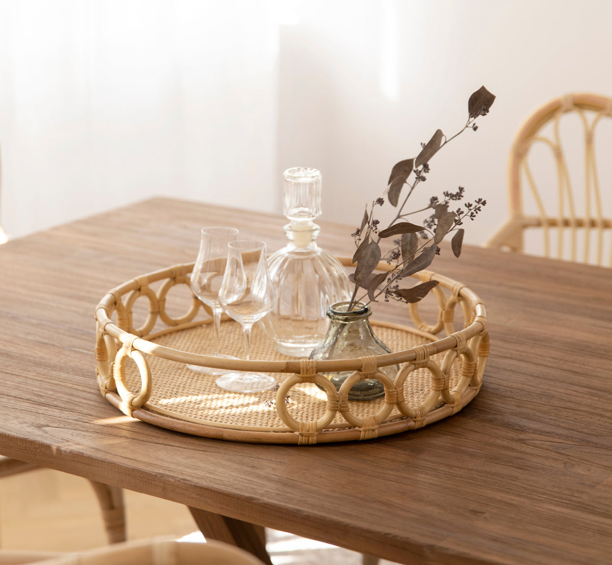 Dette er et livsstilsbillede af Mira Tray i natural rattan. På et teakbord med krystalglas og en vase med tørrede grene.